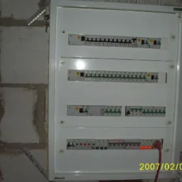 Otwarta skrzynka rozdzielcza z wieloma bezpiecznikami i wyłącznikami, zamontowana na surowej ścianie z bloków betonowych, widoczne czerwone diody LED sygnalizujące stan obwodu.