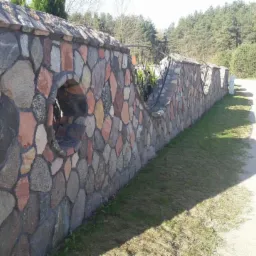 Kamienny mur ogrodzeniowy z okrągłymi prześwitami, wykonany z różnokolorowych kamieni, przylegający do trawnika i ścieżki.