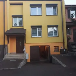 Żółta, dwupiętrowa elewacja domu z brązowymi roletami, otwartym garażem i schodami wejściowymi z metalową barierką.