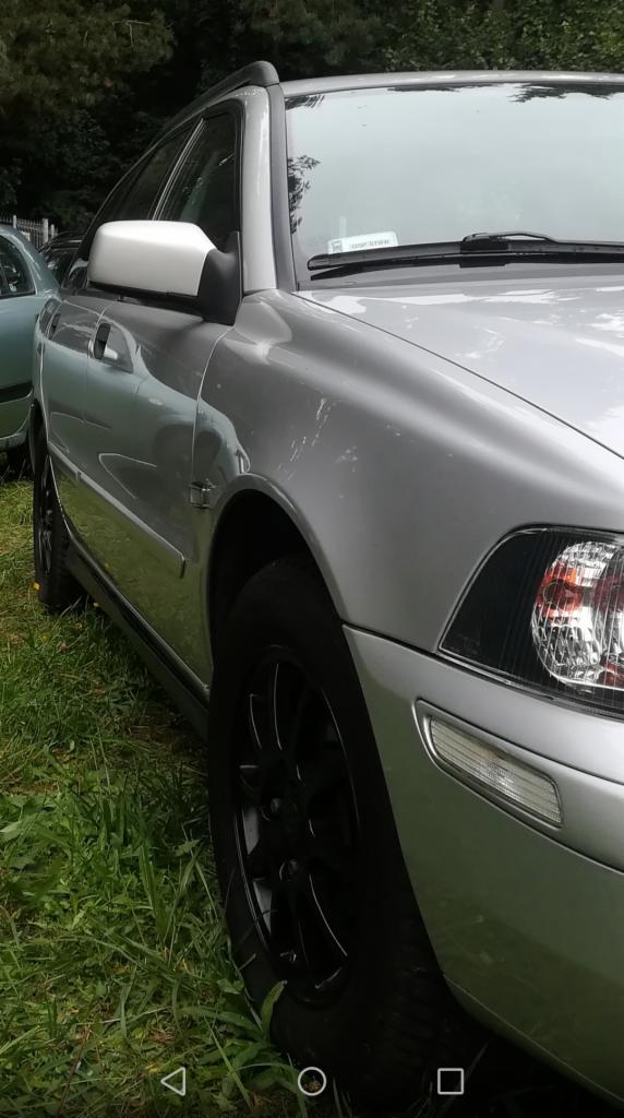 Srebrny samochód osobowy typu kombi z czarnymi felgami, zaparkowany na trawie, widoczny z perspektywy przedniego koła i reflektora.