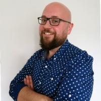 Uśmiechnięty mężczyzna w okularach i koszuli w kropki, z brodą, pozujący z założonymi rękami na tle jasnej ściany.