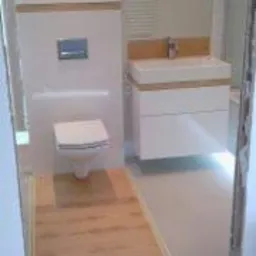 Nowoczesna łazienka z białymi meblami i drewnianymi akcentami: podłoga imitująca drewno, zabudowa toalety i umywalki, widok z perspektywy wejścia, niedokończone wykończenie.