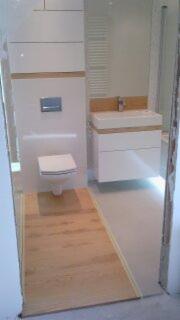 Nowoczesna łazienka z białymi meblami i drewnianymi akcentami: podłoga imitująca drewno, zabudowa toalety i umywalki, widok z perspektywy wejścia, niedokończone wykończenie.