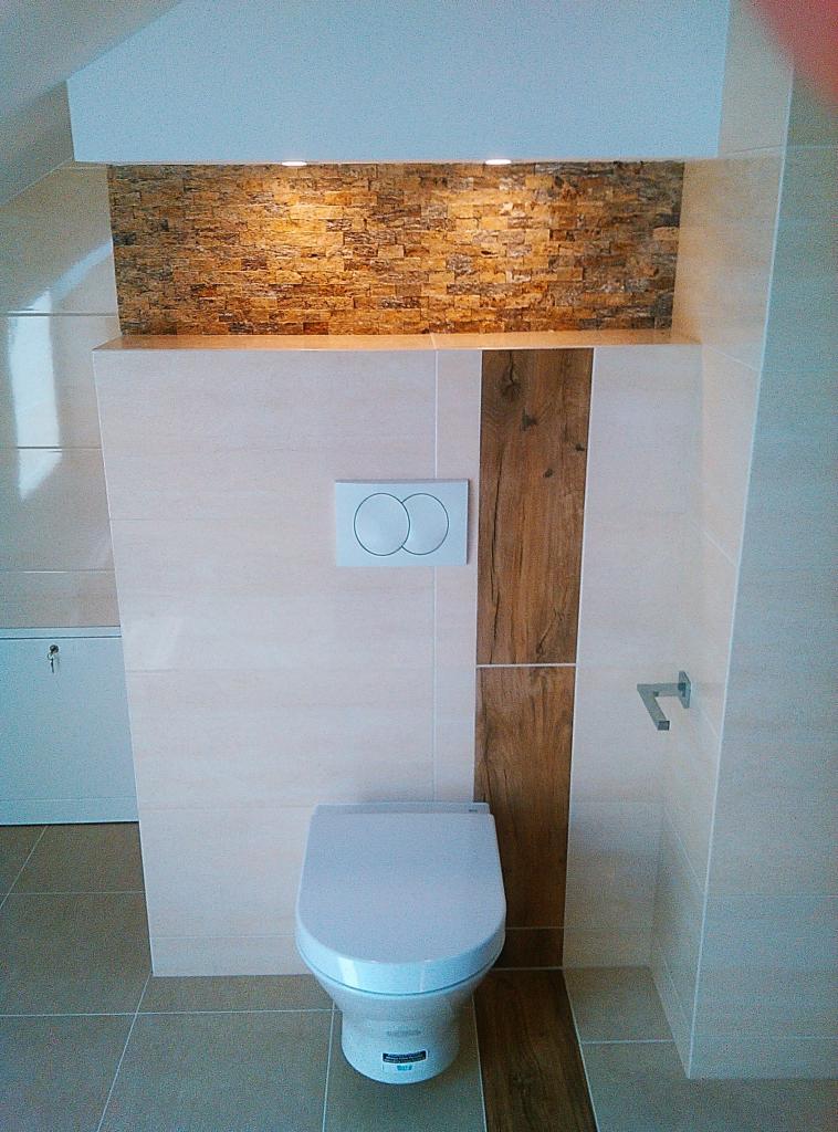 Nowoczesna toaleta podwieszana z białym przyciskiem spłukiwania, obudowana płytkami imitującymi drewno i kamień, oświetlenie punktowe nad kamienną mozaiką.