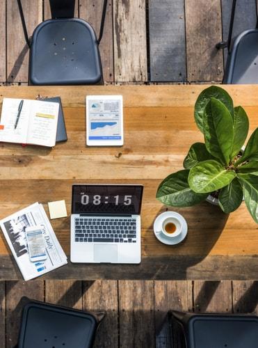 Widok z góry na biurko z laptopem, tabletem, notatnikiem, gazetą i filiżanką kawy, otoczone drewnianym blatem i krzesłami, w jasnym świetle.