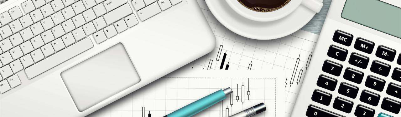 Widok z góry na biurko z białym laptopem, kalkulatorem, długopisami, filiżanką kawy i wydrukowanym wykresem giełdowym.