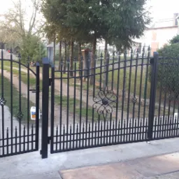 Otwarta, czarna, metalowa brama wjazdowa z ozdobnymi elementami na tle domu i drzew