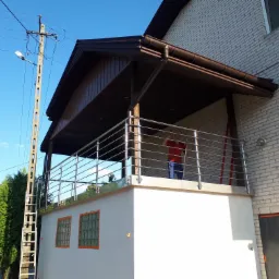 Balkon z balustradą ze stali nierdzewnej, zadaszony, z widocznym mężczyzną w czerwonej koszulce i niebieskich spodniach, biała ściana z luksferami w pomarańczowych ramach, obok słup elektryczny...