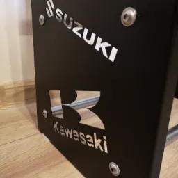 Czarny, metalowy element konstrukcyjny z wyciętymi laserowo logotypami Suzuki i Kawasaki, przymocowany śrubami, stojący na drewnianej podłodze.