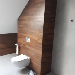 Nowoczesna łazienka z wiszącą toaletą i ścianą wykończoną panelami imitującymi drewno, widoczna instalacja podtynkową, szary gres na podłodze.