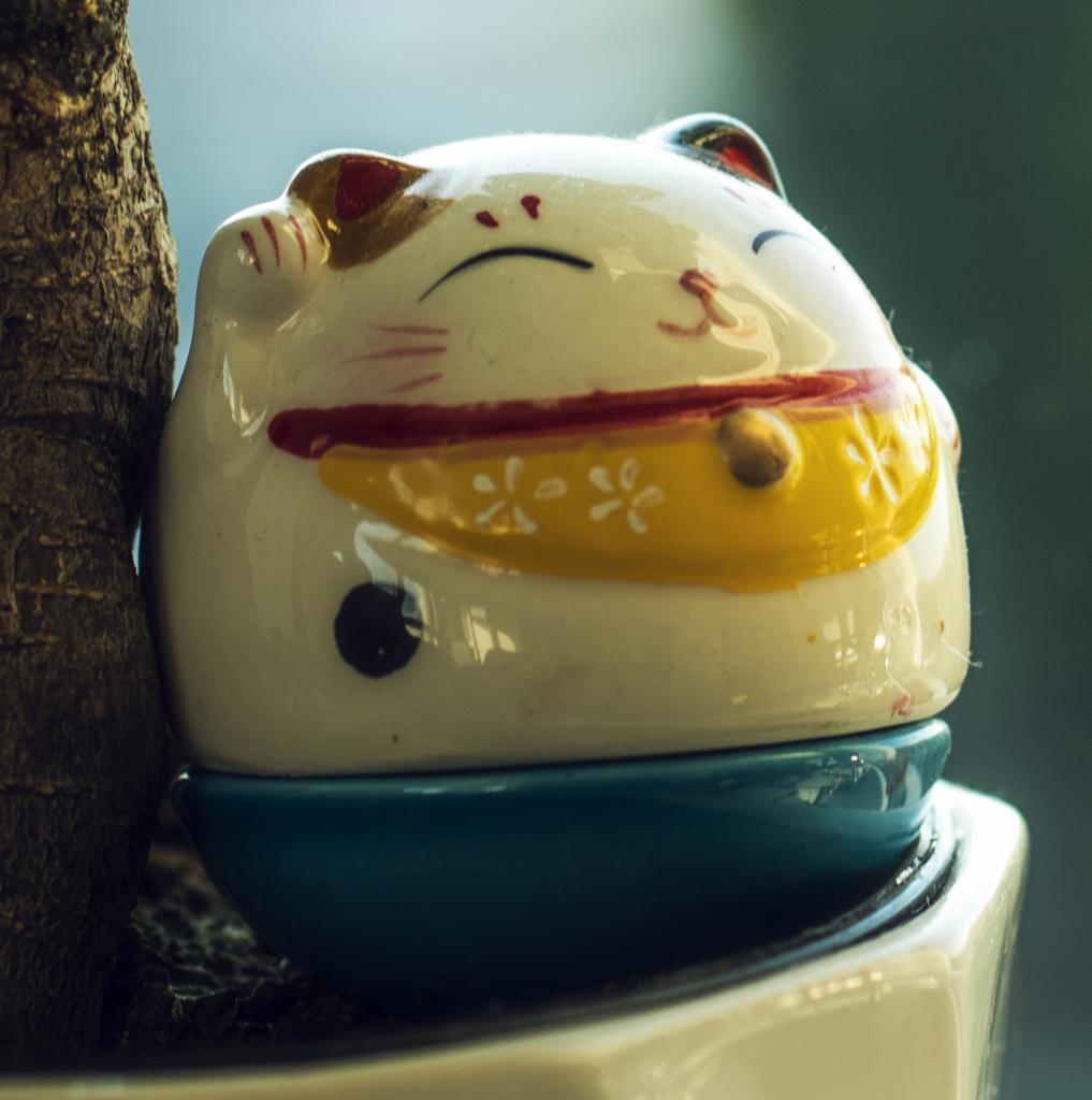Ceramiczna figurka kota Maneki Neko z podniesioną łapką, zdobiona czerwonymi i żółtymi akcentami, umieszczona na niebieskiej podstawce w białej doniczce, obok pnia drzewa.