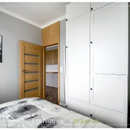 Biała szafa na wymiar z czarnymi gałkami w sypialni, widok na otwarte drzwi do kuchni, łóżko z motywem kwiatowym i plakat z górskim krajobrazem na szarej ścianie.