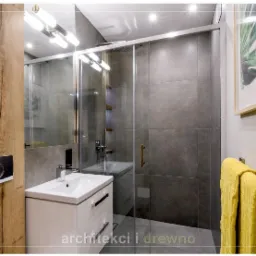 Nowoczesna łazienka z kabiną prysznicową w szarych płytkach, białą umywalką z szafką, drewnianą okładziną z przyciskiem spłuczki i żółtym ręcznikiem na wieszaku, doświetlona punktowym oświetleniem.