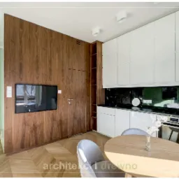 Nowoczesna kuchnia z drewnianą ścianą, ukrytymi drzwiami i telewizorem, białe szafki wiszące, czarny backsplash, okrągły stół i szare krzesła na parkiecie w jodełkę.