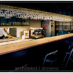 Nowoczesny bar w restauracji z drewnianym blatem, szklankami wiszącymi nad barem, profesjonalnym ekspresem do kawy i lodówką Coca-Cola, widok na pracownika za barem.