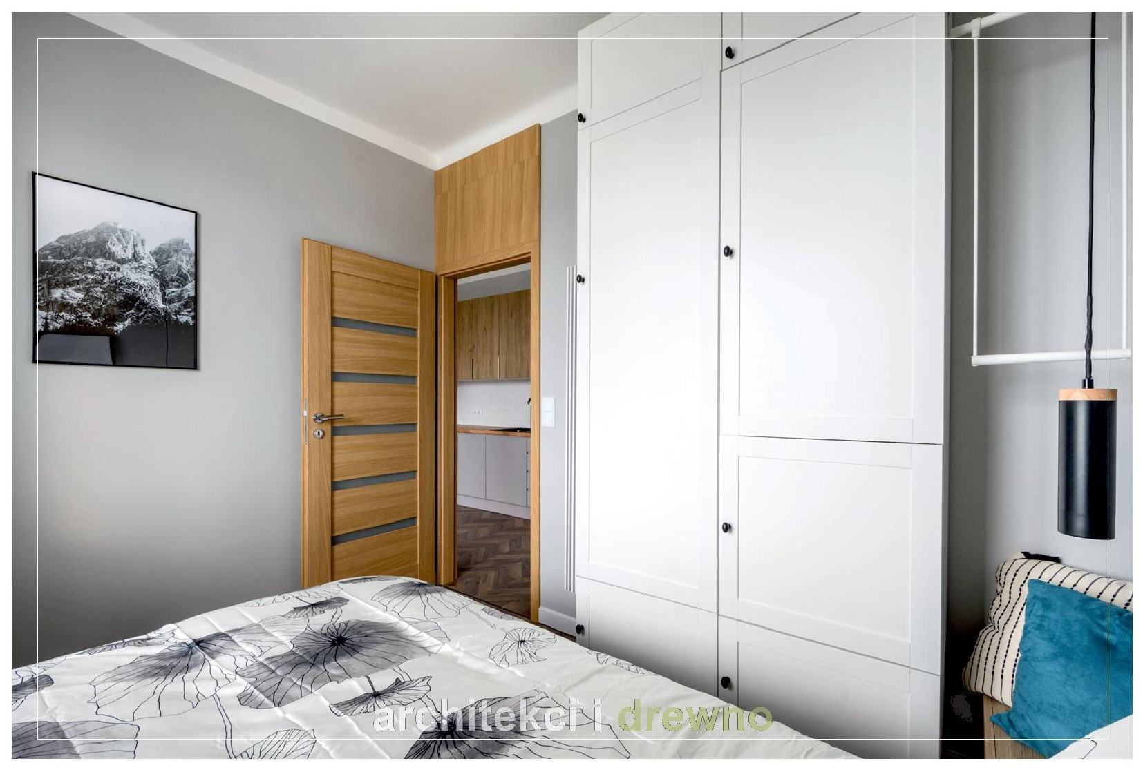 Biała szafa na wymiar z czarnymi gałkami w sypialni, widok na otwarte drzwi do kuchni, łóżko z motywem kwiatowym i plakat z górskim krajobrazem na szarej ścianie.