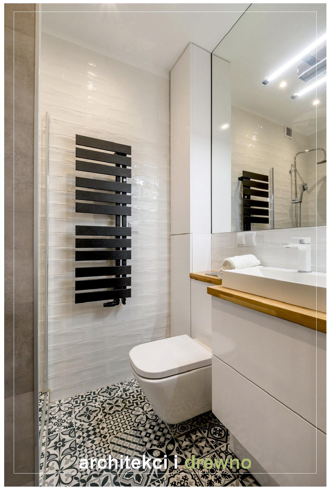 Nowoczesna łazienka z białą ceramiką, czarnym grzejnikiem drabinkowym, wzorzystą posadzką i drewnianym blatem pod umywalką.