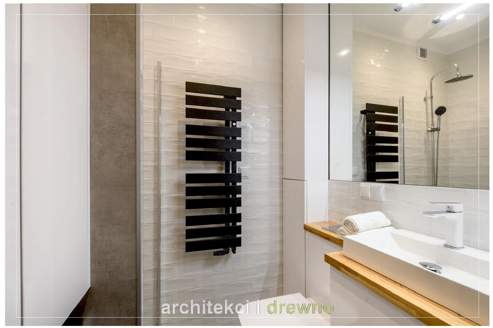 Nowoczesna łazienka z białymi płytkami imitującymi cegłę, czarnym grzejnikiem drabinkowym, białą umywalką nablatową na drewnianym blacie i dużym lustrem z odbiciem kabiny prysznicowej.