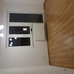 Białe okno PCV z grzejnikiem pod parapetem w pokoju z drewnianą podłogą i kinkietem na ścianie.