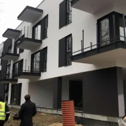 Nowoczesny budynek wielorodzinny w trakcie wykończenia, z balkonami i elewacją w kolorach bieli i grafitu, widoczni pracownicy w odblaskowych kamizelkach i rury kanalizacyjne na placu budowy.