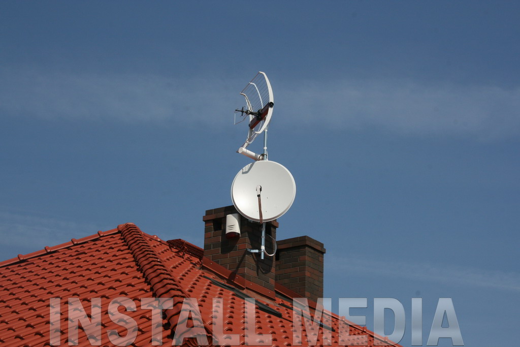 Antena satelitarna i antena kierunkowa UHF zamontowane na ceglanym kominie, widoczne na tle błękitnego nieba i czerwonego dachu z dachówki ceramicznej.