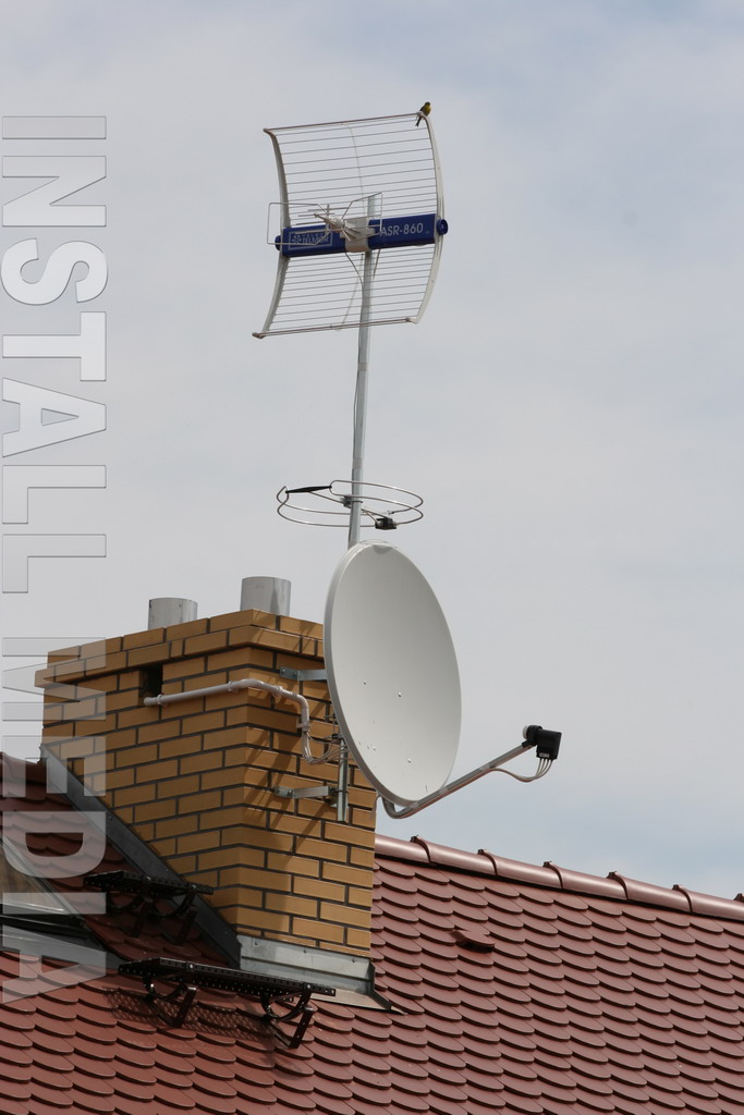 Antena telewizyjna ASR-860 i antena satelitarna zamontowane na kominie ceglanego domu z czerwoną dachówką, na antenie siedzi mały ptak.