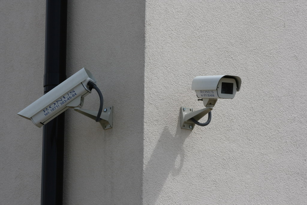 Dwie kamery monitoringu wizyjnego marki Ronus zamontowane na zewnętrznej ścianie budynku o jasnej, chropowatej fakturze, z widoczną rurą spustową obok jednej z kamer.