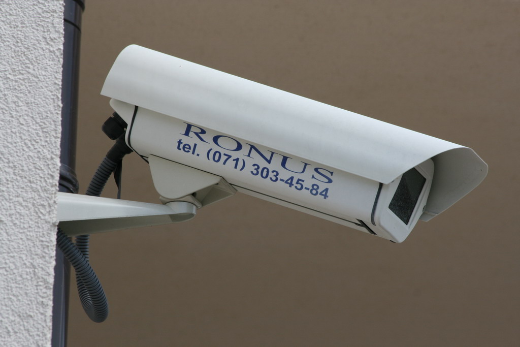 Biała kamera monitoringu z napisem 'RONUS tel. (071) 303-45-84', zamontowana na białej ścianie budynku z widoczną szarą rurą kablową.