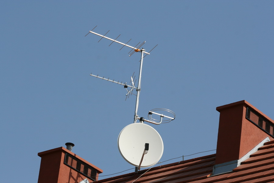Antena satelitarna i anteny naziemne zamontowane na dachu budynku z czerwonej dachówki i dwoma kominami z kratkami wentylacyjnymi, na tle błękitnego nieba.