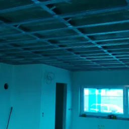 Metalowa konstrukcja sufitu podwieszanego z płyt kartonowo-gipsowych w trakcie montażu, widoczne profile i płyty, w tle okno i przygotowane materiały budowlane.