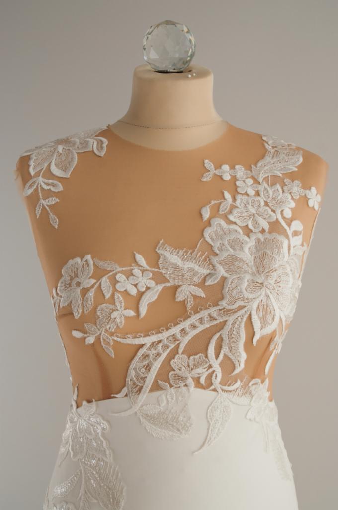 Fragment sukni ślubnej z koronkowymi aplikacjami kwiatowymi na bezbarwnym tiulu, prezentowany na manekinie z kryształową gałką.