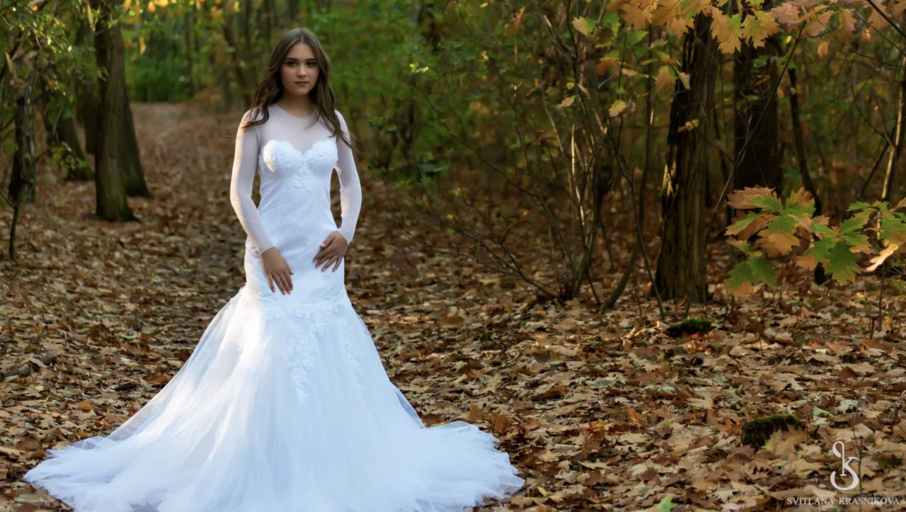 Kobieta w białej sukni ślubnej z długimi rękawami, pozująca w jesiennym lesie, z opadłymi liśćmi na ziemi.