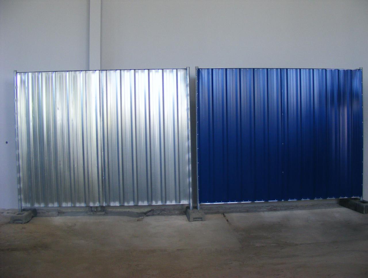 Tymczasowe ogrodzenie budowlane z blachy falistej: ocynkowana po lewej, niebieska po prawej, zamocowane na betonowych podstawach w surowym wnętrzu hali.