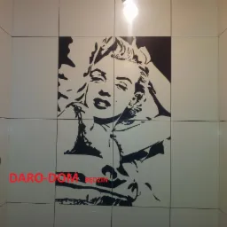 Ściana łazienki wyłożona białymi płytkami z czarnym wzorem Marilyn Monroe, oświetlona żarówką, widoczny fragment logo firmy.