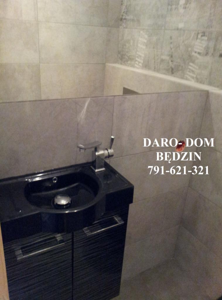 Wykończona łazienka z czarną szafką pod umywalkę, chromowaną baterią i beżowymi płytkami na ścianach, widoczne logo firmy na ścianie.