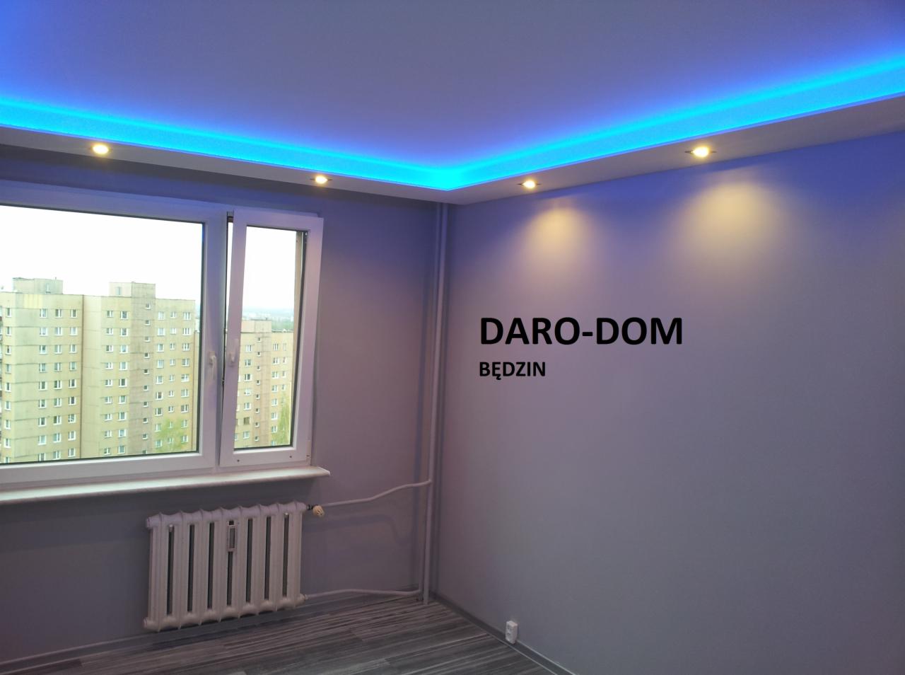 Szare wnętrze pokoju po odświeżeniu, z podwieszanym sufitem z niebieskim oświetleniem LED, widok na blok z okna, grzejnik pod oknem, na ścianie napis DARO-DOM BĘDZIN.