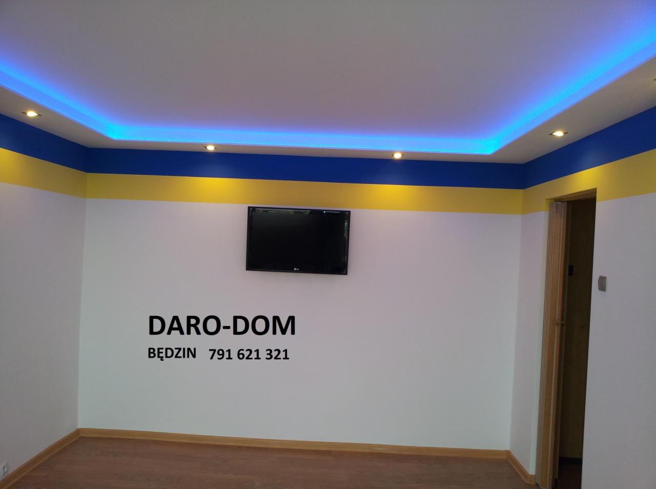 Wnętrze pokoju po malowaniu ścian na biało z poziomym pasem żółtym i niebieskim pod sufitem, podwieszany sufit z niebieskim oświetleniem LED, telewizor na ścianie, widoczne logo firmy Daro-Dom