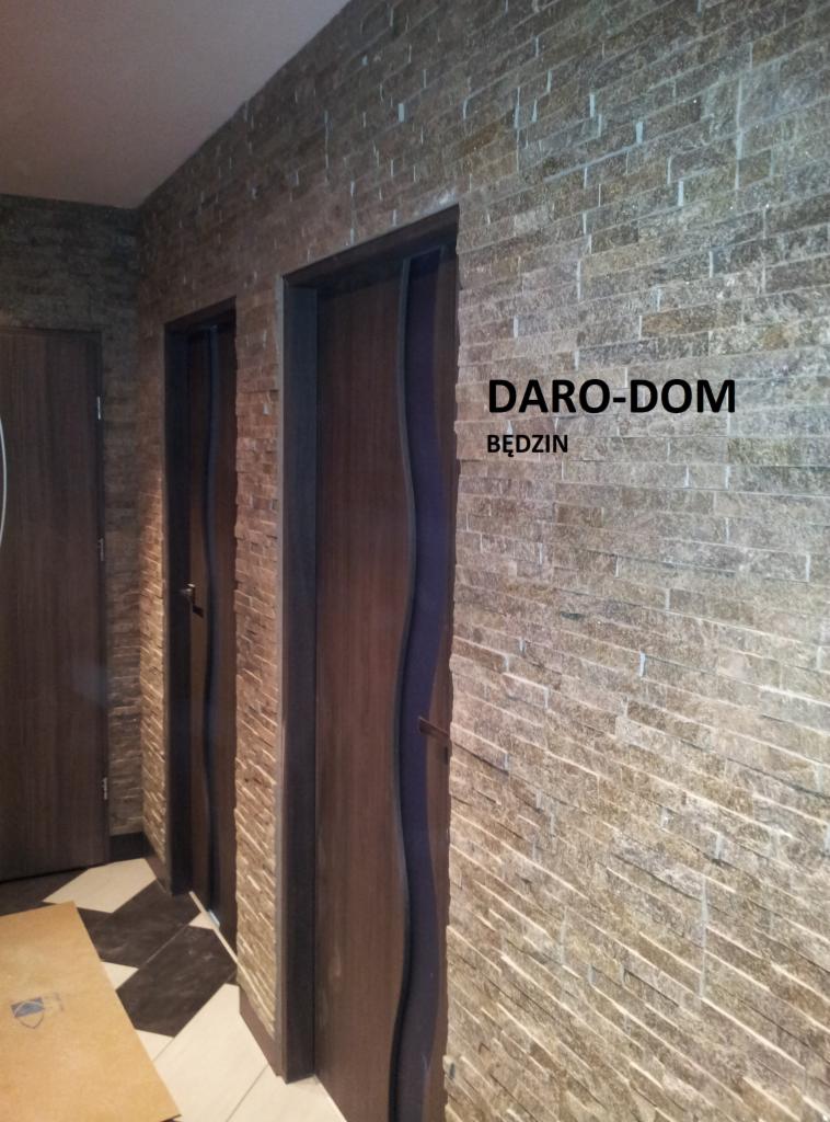 Ściana wyłożona dekoracyjną cegłą w odcieniach beżu i brązu, z widocznymi dwoma parami drzwi z ciemnego drewna z falistymi ościeżnicami, fragment podłogi z czarno-białą szachownicą. Na ścianie...