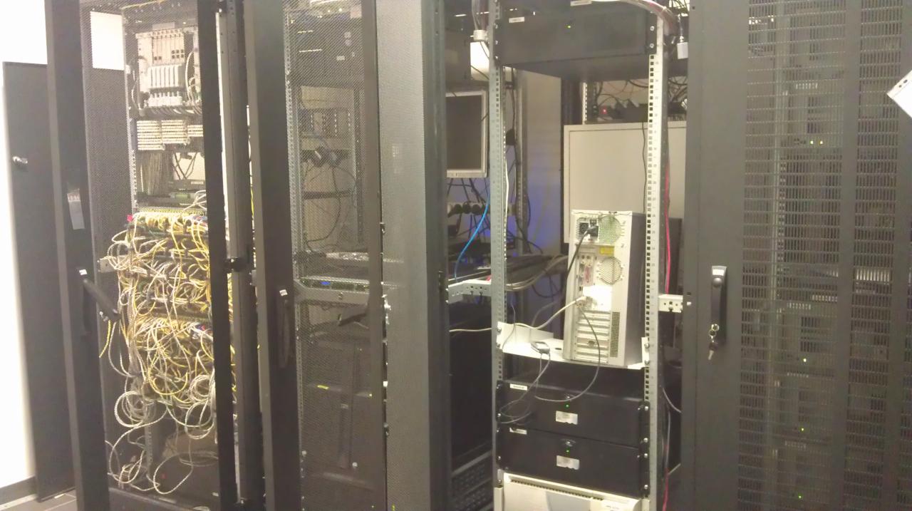 Serwerownia z szafami rack, widoczne kable i sprzęt sieciowy, w tle monitor i obudowa komputera.