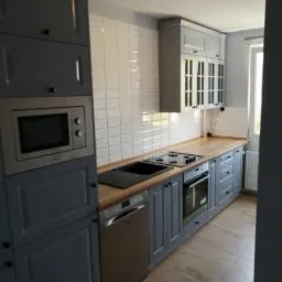 Szara zabudowa kuchenna z drewnianym blatem, białymi kafelkami i sprzętem AGD, widok z boku na gotową realizację.