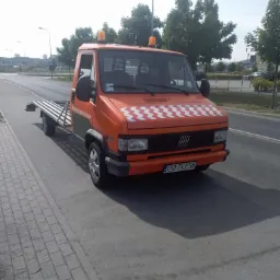 Pomarańczowa laweta Fiat Ducato z biało-czerwonym wzorem szachownicy na masce, zaparkowana na poboczu drogi w słoneczny dzień.
