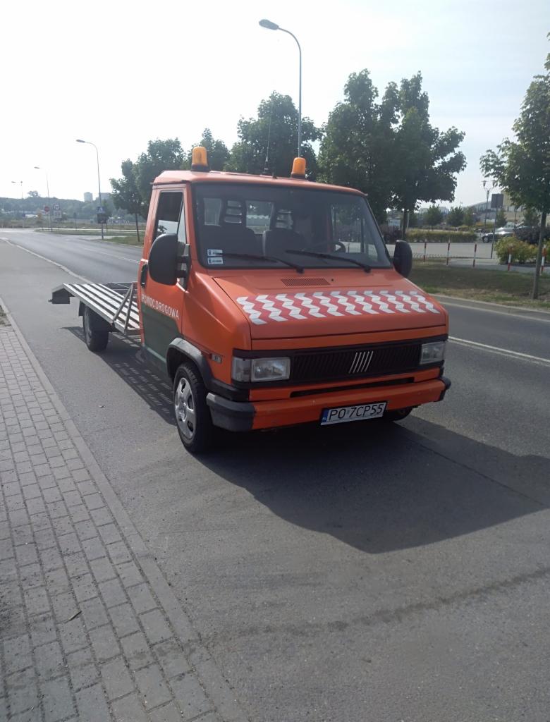 Pomarańczowa laweta Fiat Ducato z biało-czerwonym wzorem szachownicy na masce, zaparkowana na poboczu drogi w słoneczny dzień.