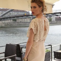 Kobieta w beżowej sukience z koronkowym topem i odkrytymi plecami, spoglądająca przez ramię na tle rzeki i mostu miejskiego.