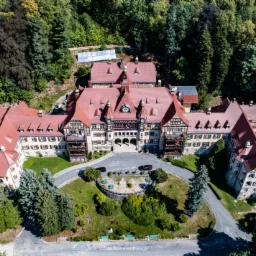 Rozległy budynek w stylu rustykalnym z czerwonym dachem, otoczony gęstym lasem, sfotografowany z lotu ptaka w słoneczny dzień. Widoczne detale architektoniczne, zadbany trawnik i zaparkowane...