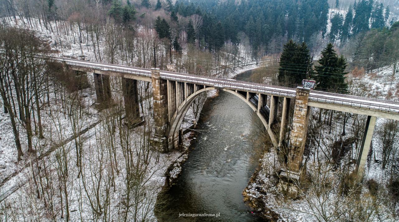 Betonowy wiadukt kolejowy z łukowymi przęsłami nad rzeką w zimowej scenerii, widok z lotu ptaka.