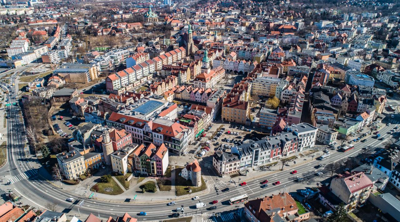 Panorama miasta z lotu ptaka, widoczne kolorowe kamienice z czerwonymi dachami, rynek z ratuszem, kościoły z wieżami, ulice z samochodami, drzewa bez liści, słoneczny dzień.