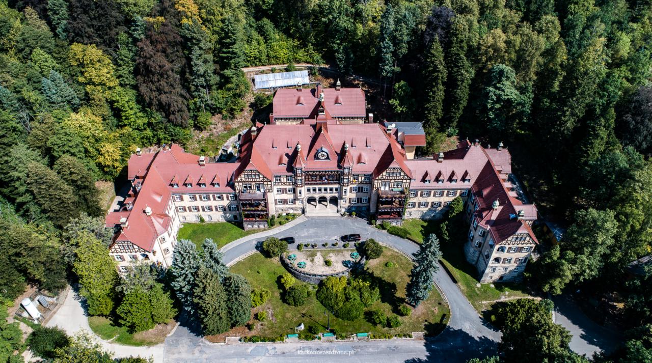 Rozległy budynek w stylu rustykalnym z czerwonym dachem, otoczony gęstym lasem, sfotografowany z lotu ptaka w słoneczny dzień. Widoczne detale architektoniczne, zadbany trawnik i zaparkowane...