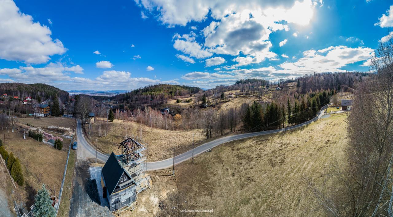 Szeroki kadr na krajobraz górski z widokiem na budowę domu z szkieletową konstrukcją, błękitne niebo z chmurami i w oddali zabudowania miejscowości.