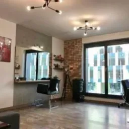 Wnętrze salonu z dwoma stanowiskami, lustrami, ceglaną ścianą i nowoczesnym oświetleniem sufitowym. Na ścianach wiszą obrazy. Widoczny parkiet i duże okno.