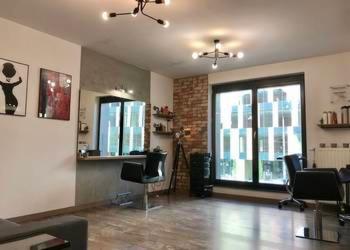 Wnętrze salonu z dwoma stanowiskami, lustrami, ceglaną ścianą i nowoczesnym oświetleniem sufitowym. Na ścianach wiszą obrazy. Widoczny parkiet i duże okno.